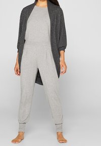 Tuta grigia con una vestibilità rilassata e pieghe laterali, abbinata a un cardigan aperto grigio scuro e texturizzato. Entrambi hanno maniche lunghe.