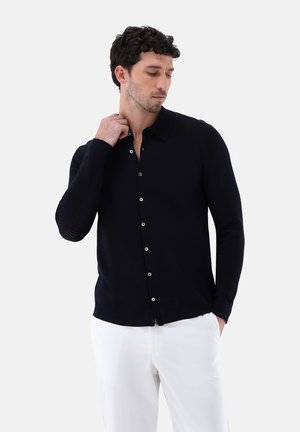 Marineblaues, gestricktes Button-up-Hemd mit spitzem Kragen, langen Ärmeln und glatter Textur, kombiniert mit weißen Hosen. Subtile Knopfdetails sichtbar.