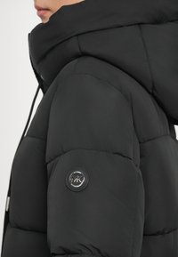 Must komforttäidisega jope suure kapuutsiga, millel on tikitud muster, varrukal ümmargune logomärk ja sile viimistlus.