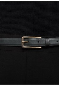 Ceinture en cuir noir avec une boucle rectangulaire argentée attachée autour d’un tissu noir tricoté et d’un pantalon noir.