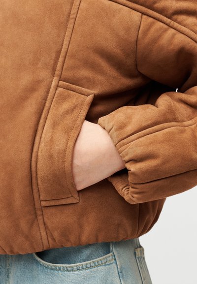 Veste en suédine marron à la texture douce, avec une poche latérale et des poignets élastiques, présentée en gros plan sur un denim bleu clair.