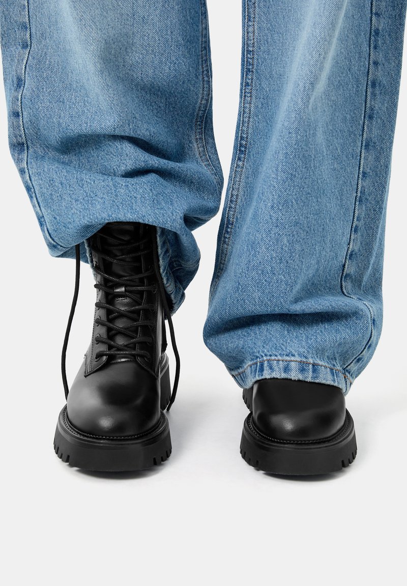 Bottes noires à lacets avec un bout arrondi, semelle en caoutchouc épaisse, associées à un jean en denim bleu clair ample.
