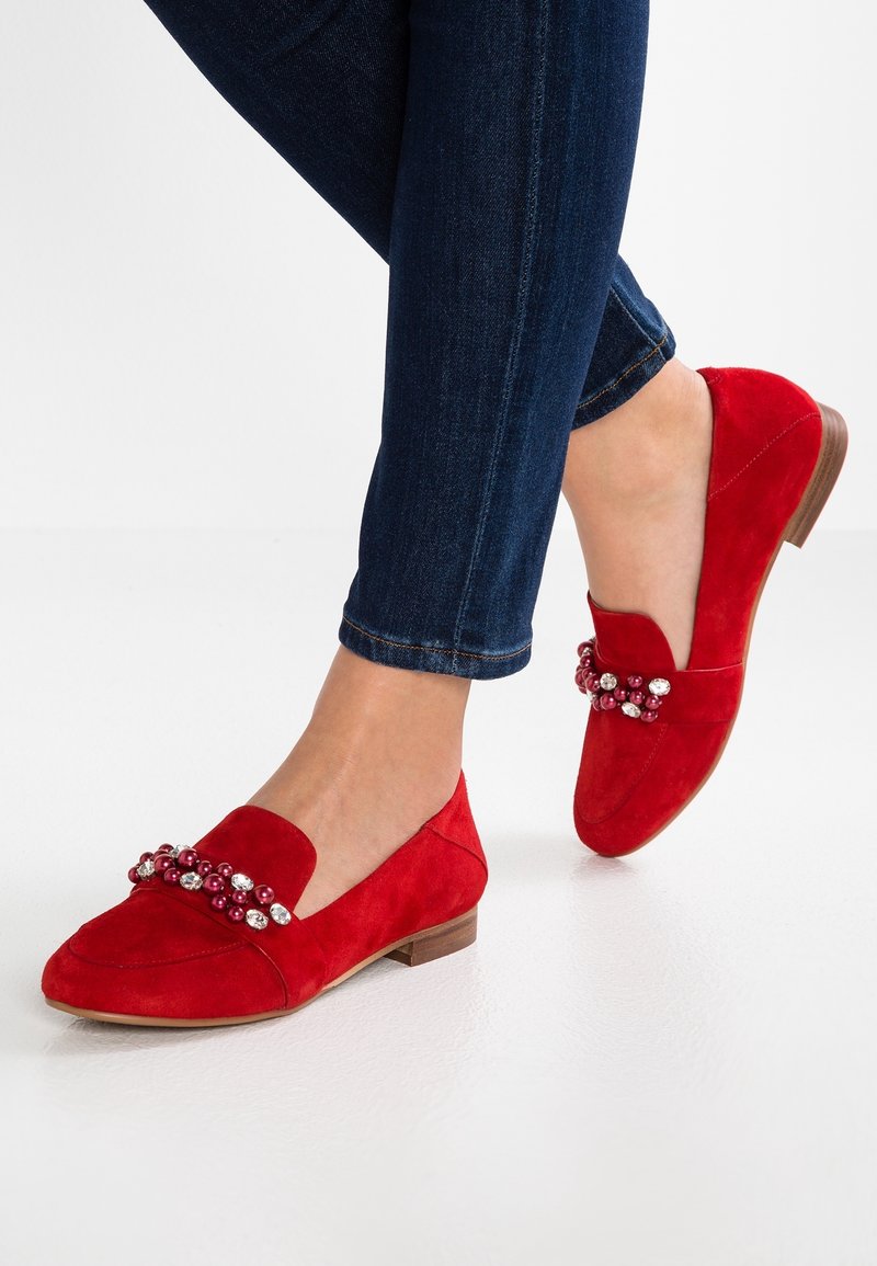 Mocassins en daim rouge avec des embellissements perlés portés avec un jean skinny bleu foncé sur fond blanc.