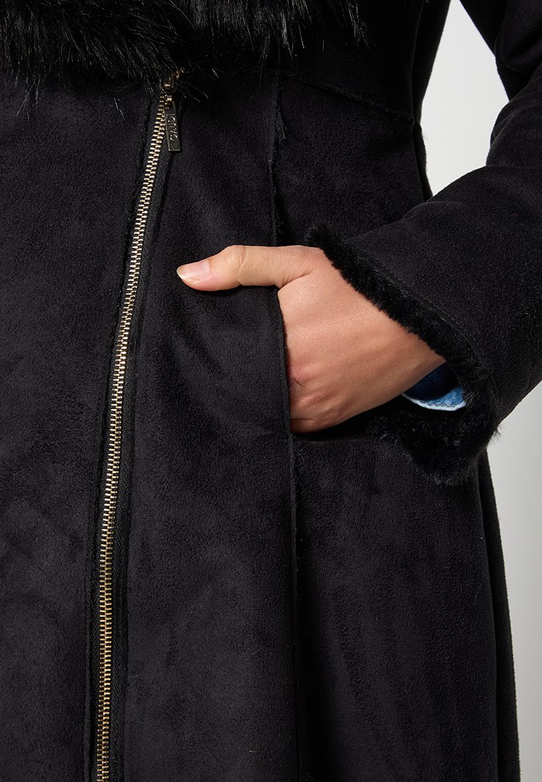 Cappotto in finta suede nero con colletto in pelliccia morbida, dotato di zip dorata e tasca laterale. Texture liscia con leggero lucido.