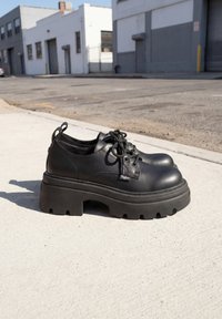 Paar zwarte leren platform veterschoenen met dikke zolen geplaatst op een betonnen stoep nabij een industriële straat.