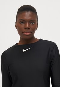 Juodas ilgomis rankovėmis megztinis, pagamintas iš lygaus audinio, su baltu Nike logotipu ant krūtinės. Paprastas apvalus iškirpimas ir prigludusi forma.