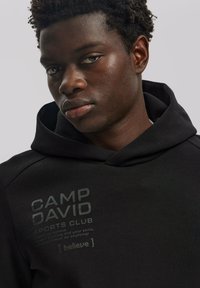 Fekete kapucnis felső texturált felülettel, elöl szürke színben nyomtatott "CAMP DAVID SPORTS CLUB" logóval és felirattal. Egyszerű, sportos dizájn.