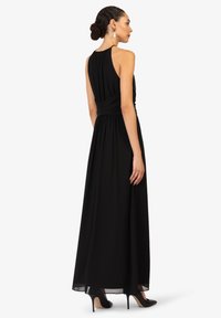Schwarzes Maxikleid mit hohem Kragen, gerafftem Oberteil und fließendem Rock. Es hat eine figurbetonte Taille und einen glatten Stoff. Trägt schwarze High Heels.