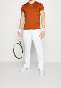 Tennisoutfit med en brun kortärmad skjorta, vita lösa byxor och vita sneakers, håller en svart tennisracket.