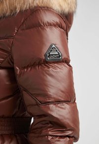 Brun puffjacka med en glänsande yta, med en triangel-logotyp på ärmen märkt "JACKIT". Fuskpälskrage accentuerar designen.