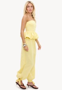 Femme blonde debout dans une combinaison jaune bustier avec taille à volants, chevilles retroussées nouées avec des nœuds, sandales noires et bracelets en métal mélangé.