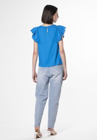 Blusa blu con maniche a rouches, caratterizzata da un dettaglio a oblò sulla schiena. Abbinata a pantaloni in denim chiaro e mule bianche con tacco. Sfondo pulito.