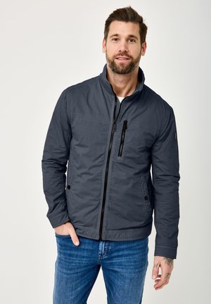 Mann mit Bart, der eine dunkelgraue Jacke mit Reißverschluss und blaue Jeans trägt, steht mit einer Hand in der Tasche vor einem einfarbigen Hintergrund.