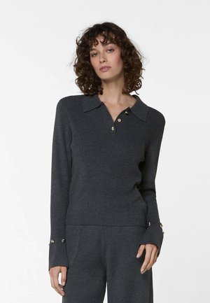 Femme portant un pull en maille côtelée gris foncé avec des détails de boutons dorés sur le col et les poignets, assorti à un pantalon coordonné.