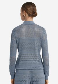Ljusblå stickad tröja med mönstrade meshsektioner, ribbade muddar och krage. Bakvy som framhäver detaljerad textur och design.