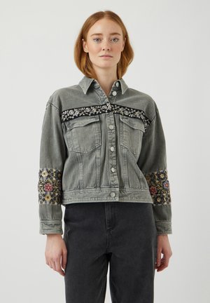 Junge Frau trägt eine graue Jeansjacke mit floral bestickten Bändern über Brust und Ärmeln, kombiniert mit dunkler Hose vor einfarbigem Hintergrund.