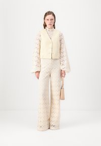 Outfit color crema composto da una maglietta in pizzo texturizzato a maniche lunghe, un gilet corto e pantaloni a gamba larga con motivo. Gli accessori includono una piccola borsa.