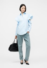 Femme portant une chemise bleu clair à manches à volants, un jean bleu clair, des talons noirs et tenant un sac à main noir.