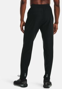 Bakifrån av en person som bär svarta träningsbyxor och svarta löparskor med reflekterande detaljer mot en vit bakgrund.