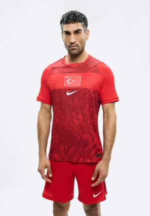 Mand iført rød sportstrøje med tyrkisk flag og Nike-logo på skjorten og Nike-logo på shorts, stående mod en hvid baggrund.