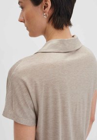 Polo shirt beige a maglia con colletto, maniche corte e una trama sottile. Orecchino a cerchio d'argento visibile nell'orecchio del modello.