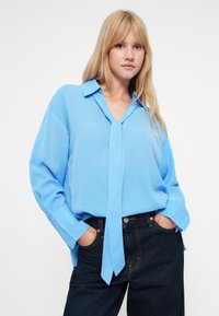 Απαλή γαλάζια μπλούζα φτιαγμένη από μαλακό, λείο ύφασμα, με γιακά, μακριά μανίκια και λεπτομέρεια με κομπίνα στον λαιμό. Συνδυάζεται με σκούρα τζιν.