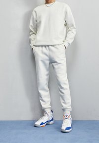 Man i en vit sweatshirt och joggers med högskor i blått, vitt och orange som står mot en ljusgrå vägg.