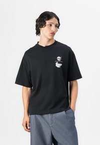 Black cotton t-shirt with short sleeves, featuring a graphic pocket design and a round neckline, worn with light blue wide-leg pants.