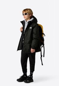 Black waterproof jacket with a hood, gray sweatshirt, black pants, and black sneakers. Wearing sunglasses, holding a yellow backpack.