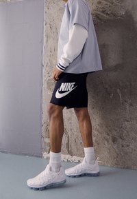 Vita Nike-sneakers med genomskinliga såler, svarta shorts med en stor vit Nike-logga, och en ljusblå oversized skjorta.