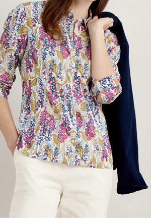 Kobieta w kwiecistej koszuli na guziki z podwiniętymi rękawami, białych spodniach, trzymająca ciemną kurtkę na ramieniu.