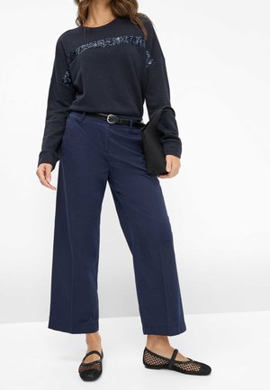 Femme portant un pantalon large bleu marine, un sweat-shirt bleu marine foncé avec une bande de sequins, une ceinture noire, des ballerines en mesh noir et tenant un sac à main noir.