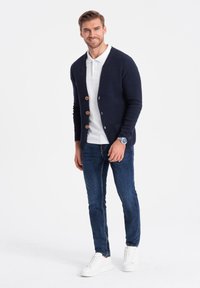Cárdigan navy de punto texturizado, con tres botones de madera, combinado con una camiseta polo blanca, jeans azul oscuro y zapatillas blancas.