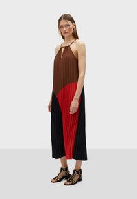 Vestito plissettato marrone e rosso con design senza maniche, caratterizzato da un collo a chiave e dettagli neri sul fondo. Include sandali neri con borchie.