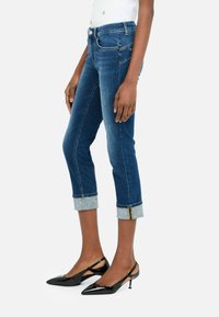 Jean en denim bleu avec un design ajusté, une longueur courte et des ourlets retroussés. Les ourlets présentent un tissu bleu clair contrastant avec un motif subtil.