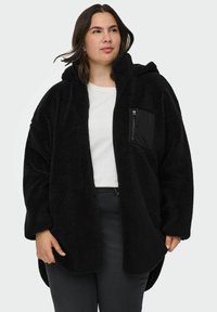 Melna vilnas jaka ar kapuci, priekšējo rāvējslēdzēju kabatu un pārspīlētu piegriezumu. Izceļas ar teksturētu, pūkainu audumu un kakla izgriezumu kā krūzes formas dizainu.