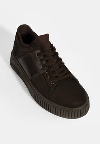 Brauner Sneaker aus strukturiertem Stoff mit glatten Akzenten, ausgestattet mit einer dicken Gummisohle und einer runden Zehenpartie. Die Schnürsenkel sind in einem Kreuzmuster geflochten.