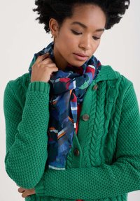 Grüne Strickjacke mit strukturiertem Muster und Knöpfen, kombiniert mit einem gemusterten Schal, der blaue, rote und grüne Akzente aufweist.