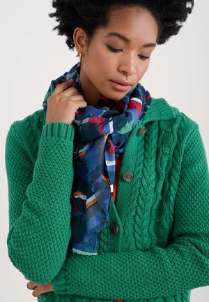 Grüne Strickjacke mit strukturiertem Muster und Knöpfen, kombiniert mit einem gemusterten Schal, der blaue, rote und grüne Akzente aufweist.
