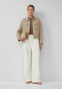 Giacca corta beige con due tasche anteriori, abbinata a un maglione marrone strutturato e pantaloni bianchi larghi. Il modello tiene una borsa crema.