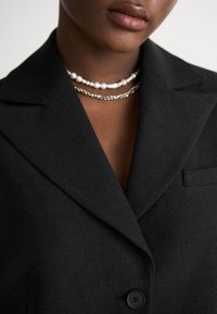 Blazer nero in tessuto texturizzato, chiusura con un solo bottone, revers a punta e vestibilità slim. Indossato con catene di perle e argento a strati.