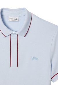 Polo Lacoste bleu clair avec des bordures rouges sur le col et la patte de boutonnage, logo crocodile bleu clair brodé sur la poitrine.
