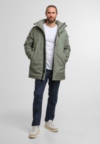 Giacca imbottita verde oliva con cappuccio, chiusura a zip e tasche frontali. Indossata sopra una camicia bianca e pantaloni blu navy. Calzature casual in beige.