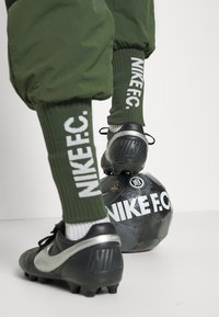 Schwarze Nike-Fußballschuhe auf einem schwarzen Nike F.C. Fußball, kombiniert mit grünen Strümpfen aus Strick mit weißem Text und lockeren grünen Jogginghosen.