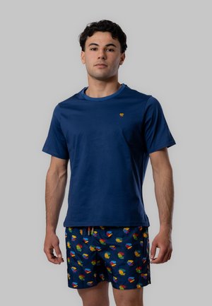 Giovane uomo in piedi che indossa una maglietta blu navy tinta unita e pantaloncini blu navy con motivi astratti colorati su sfondo grigio.