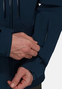 Veste marine dotée d'une poche zippée sur la manche et d'un poignet ajustable. Fabriquée en matériau lisse et durable avec un design ajusté.