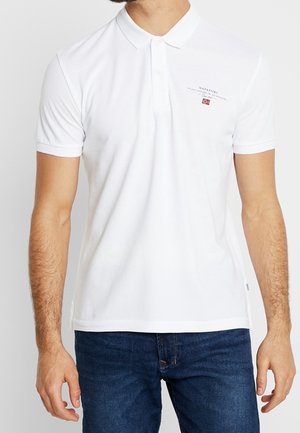 Mężczyzna noszący białą, krótką koszulkę polo z małym logo i tekstem na lewej piersi, w połączeniu z niebieskimi jeansami.