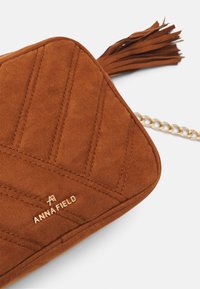Hnědá semišová taška s prošívaným designem, zlatým řetízkovým popruhem a dekorativní třásní. Na přední straně je embosované logo "ANNAFIELD".