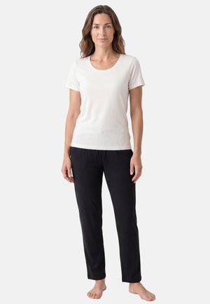Femme debout pieds nus, vêtue d'une chemise blanche à manches courtes et d'un pantalon noir devant un fond clair uni.