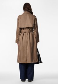 Brun trenchcoat med belte i midjen og brede slag, med delt bakdesign, kombinert med mørkeblå vide jeans og svarte sko.